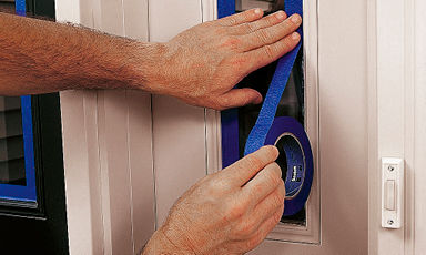 To hender fester blå maskeringstape rundt smalt vindu i beige dør ved dørklokke.