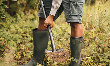 Person i grønne gummistøvler og shorts graver med spade i frodig skogbunn.