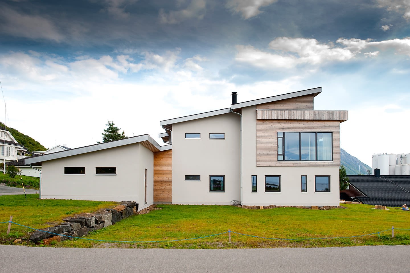 Moderne hvitt og lyst trehus med skråtak, grønt plenareal, fjell og skyet himmel.