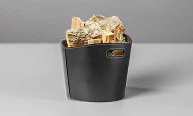 Svart lærkurv fylt med bjørkved på grå bord- og bakgrunn.