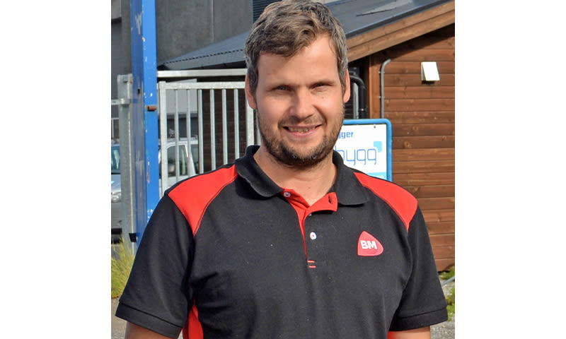 Mann i svart- og rød BM-polo utenfor byggevarebutikk med metallgjerde og trekledning.