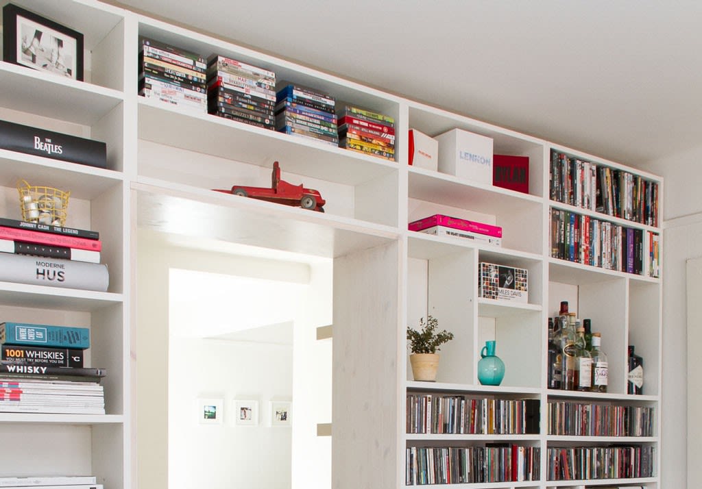 Hvit innebygd bokhylle med fargerike bøker, CD-er, DVD-er, flasker, rød lekebil og plante