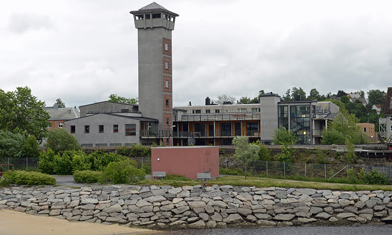 Grå betongbygning med vaktårn og glassfasade ved steinfylt elvebredd under overskyet himmel.