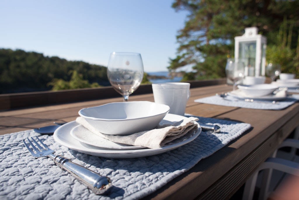 Utendørs spisebord i tre med hvite servise, glass, blå bordbrikker og sjøutsikt bak.