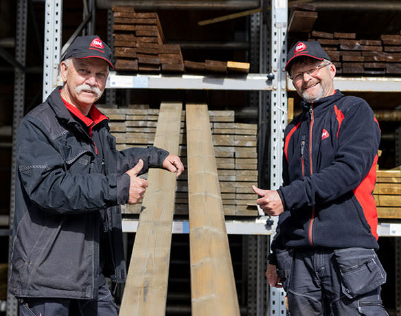 To ansatte i sort-rød arbeidsantrekk gir tommel opp ved to lange treplanker i tømmerlager.