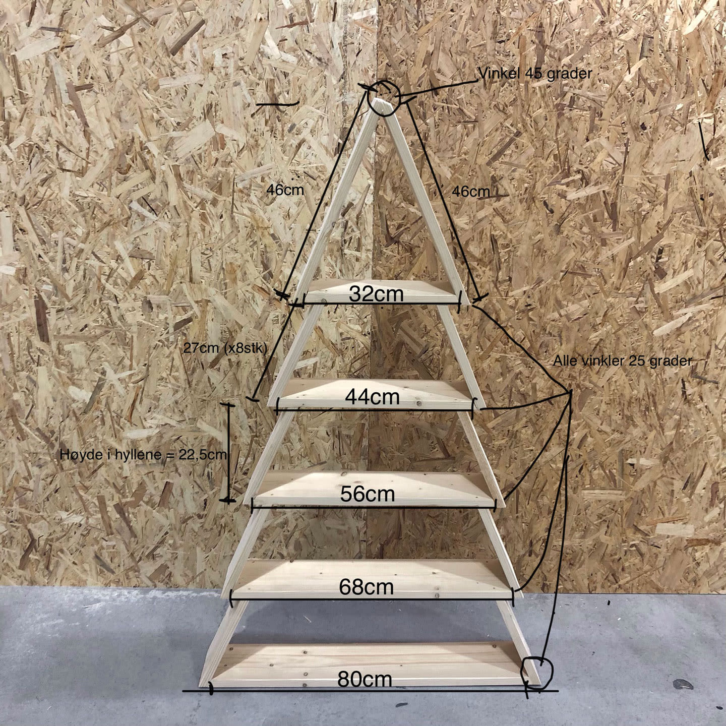 Trepyramidehylle med fem planker mot sponplatevegg på betonggulv, merket med mål.