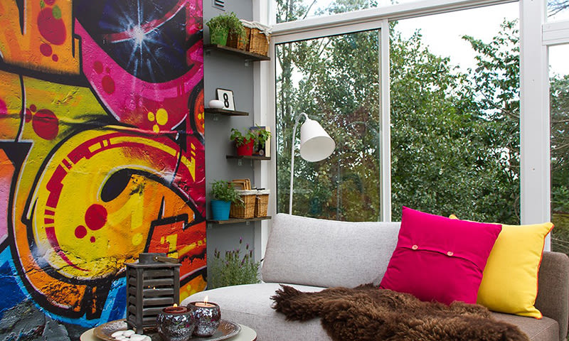 Stuekrok med grå sofa, rosa og gul pute, brunt pledd, fargerik graffiti og grønt utsyn.