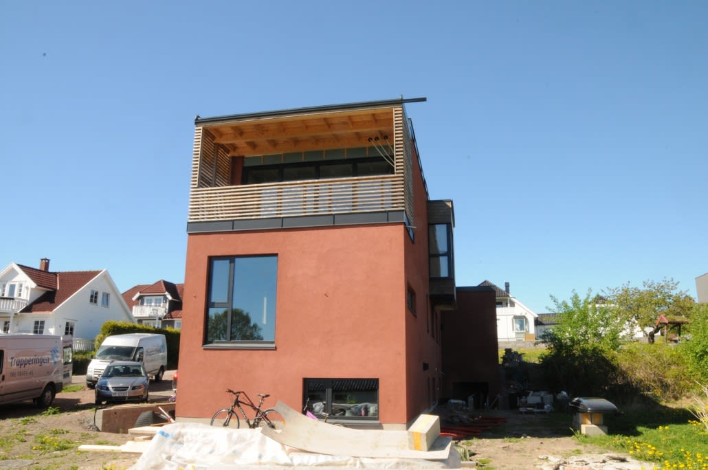 Rødt funkishus under bygging med trelamell-balkong, sykkel og blå skyfri himmel.