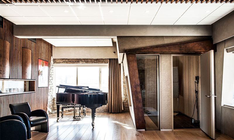 Moderne øvingsrom med svart flygel, lyse tregulv, steinvegg, sorte stoler og vokalboks.