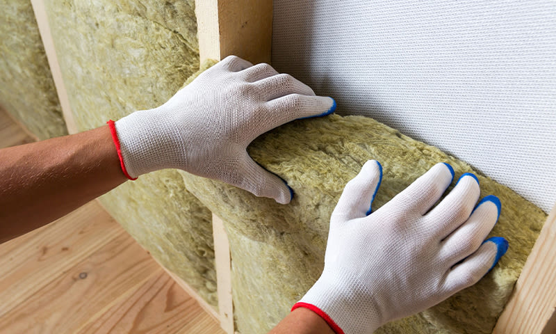 Hvite hansker med blå tupper presser gulgrønn isolasjon mellom trelekter på vegg.