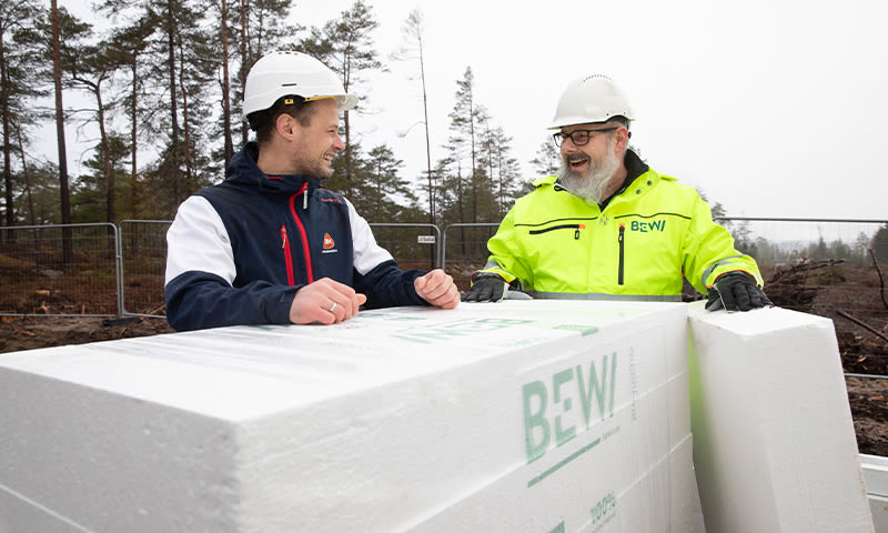 To byggarbeidere i hvite hjelmer og gul jakke ved hvite BEWI-isolasjonspaneler i skog