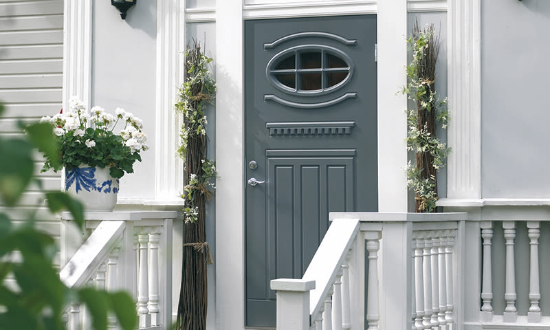 Grå dør med ovalt vindu i hvit fasade, hvit veranda med potte hvite blomster og greinbuketter