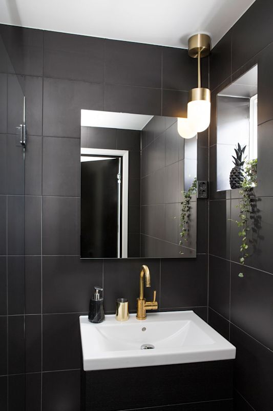 Sort flislagt baderom med hvit servant, gulldetaljer, speil, hengende lyspære og grønne planter