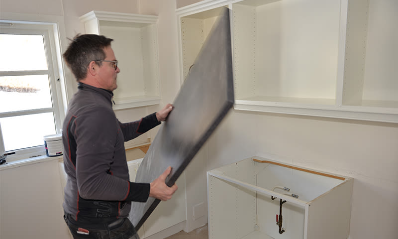 Håndverker løfter svart benkeplate i hvitt kjøkken under montering av skap