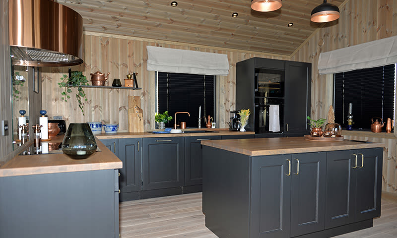 Lyst tømmerpanelert kjøkken med svarte skap, trebenk, kobberdetaljer og grønne planter