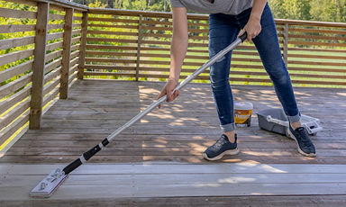 Nærbilde: person i jeans og joggesko maler terrassegulv hvitt med rull, gjerde og grønn hage