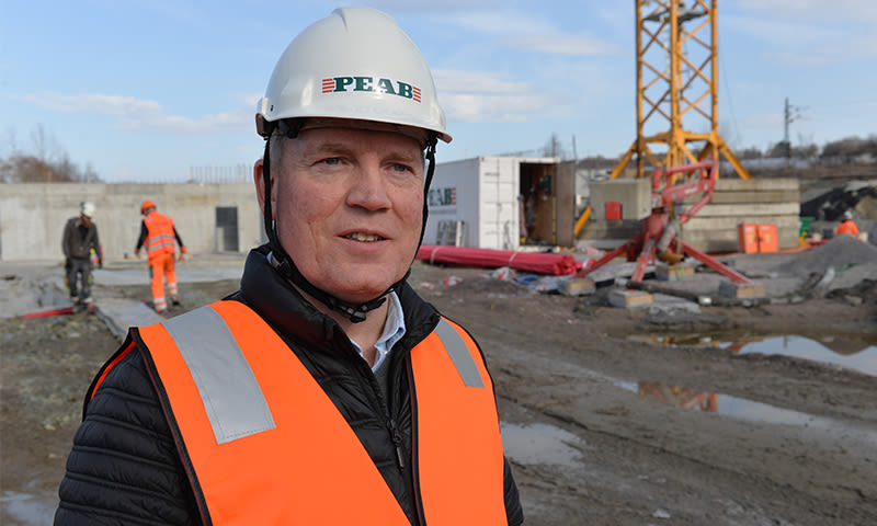 Person i hvit PEAB-hjelm og oransje vest på mudret byggeplass med kran i bakgrunnen.