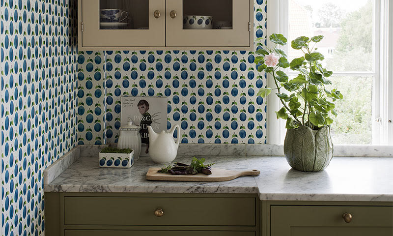 Kjøkkenhjørne med olivengrønne skap, marmorbenkeplate, pæremønstret tapet og potteplante.