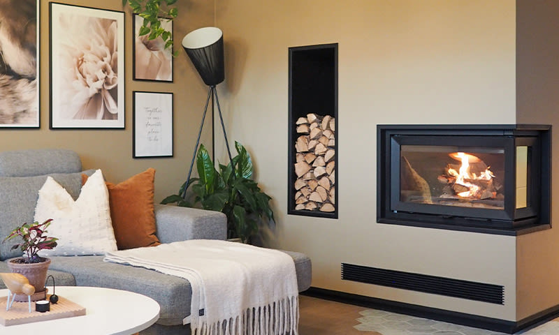 Grå sofa med hvit pledd og pute, grønne planter, svart hjørnepeis med flammer i beige stue.