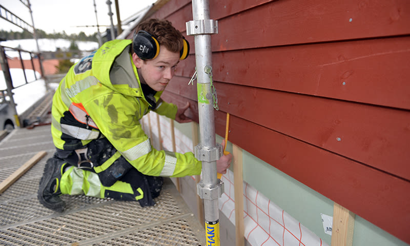 Bygningsarbeider i gul refleksjakke måler trepanel under rød tømmervegg fra stillas