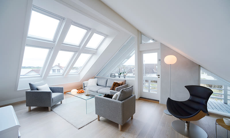 Lyst loftstue med skråtak, store takvinduer, grå sofaer, glassbord, svart lenestol