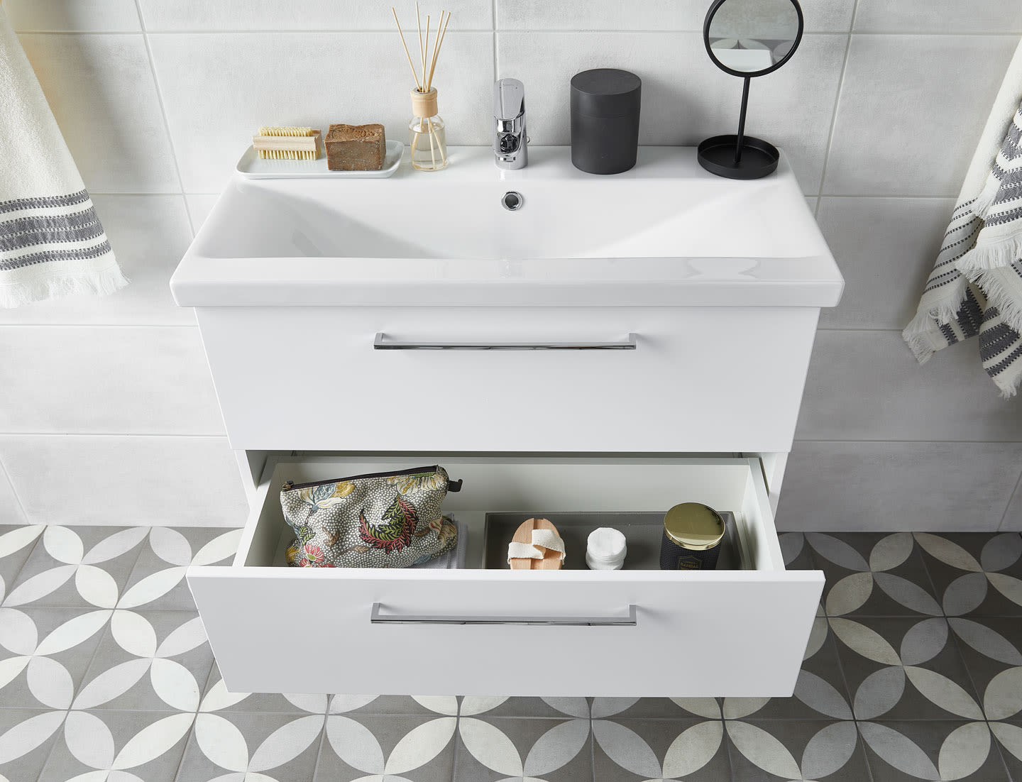 Hvit vegghengt servantskap med åpen skuff, toalettmappe, rundt speil og grått flismønster