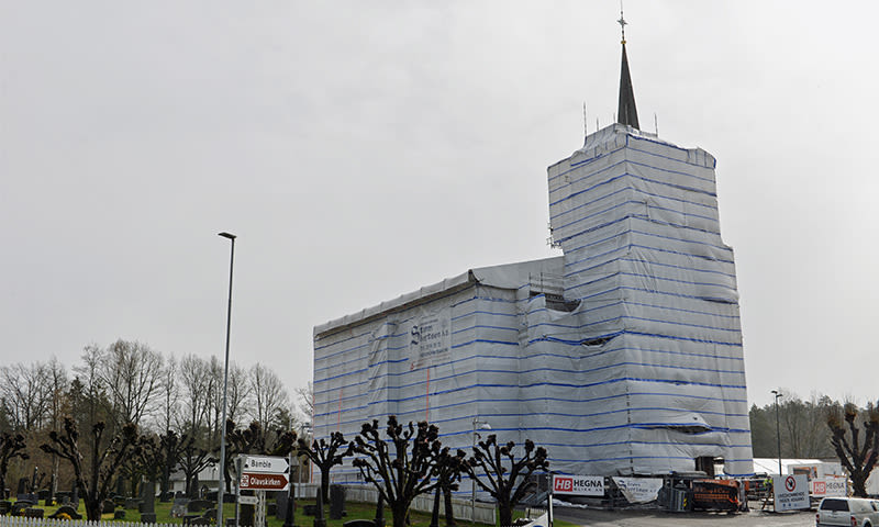 Kirke i gravlund innpakket i hvit- og blåstripet presenning, tårnspire stikker opp.