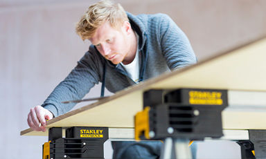 Person i grå genser måler lyst treverk på svartgul Stanley arbeidsbenk.
