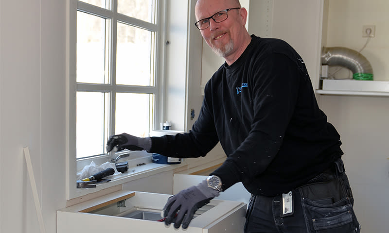 Håndverker i svart genser og grå hansker monterer hvitt kjøkkenskap under vindu.