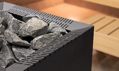 Nærbilde av svart badstuovn med grå steiner og lys trebenk i bakgrunnen.