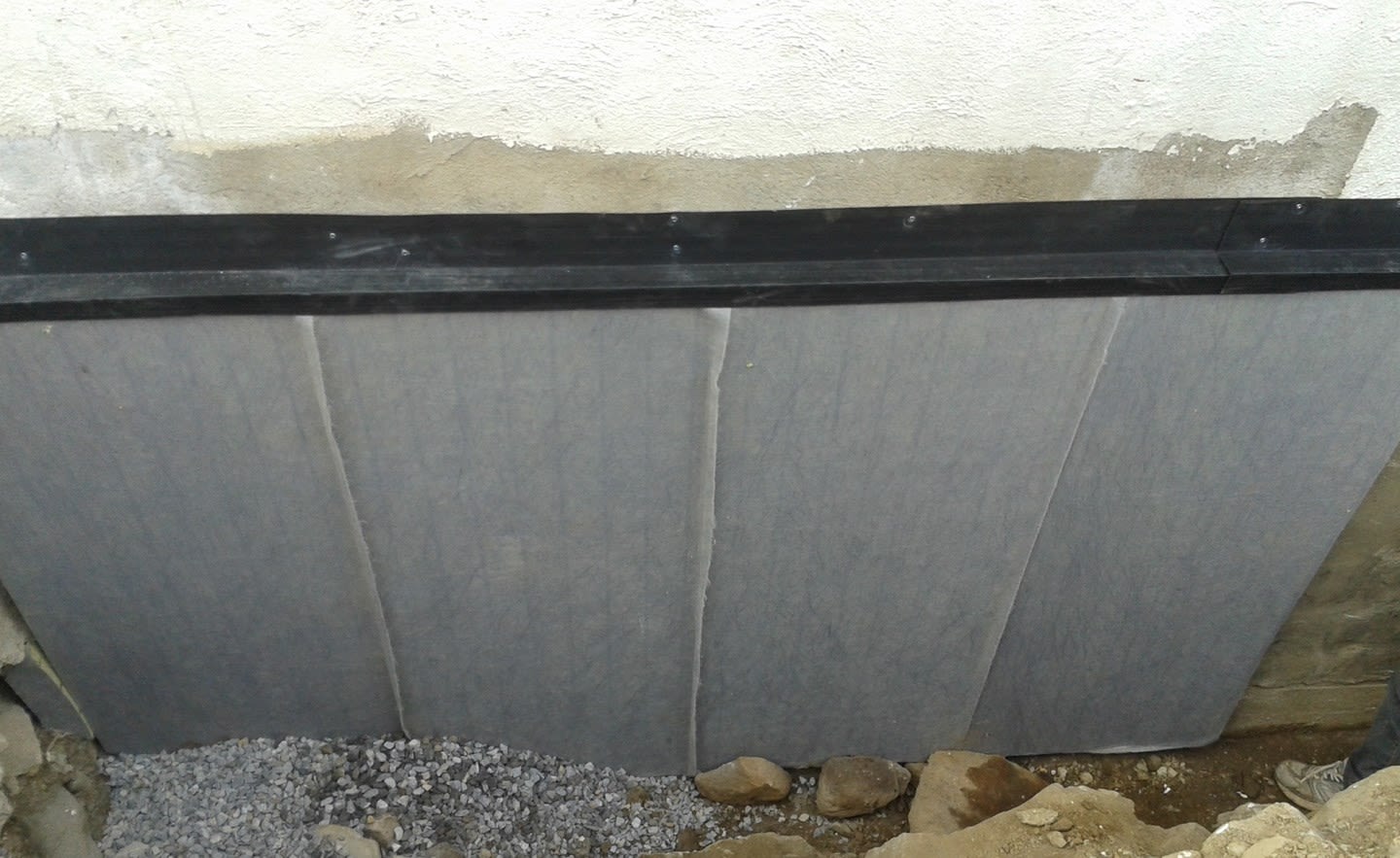 Lysegrå dreneringsplater mot hvit grunnmur, svart topplist og grå pukk nederst.
