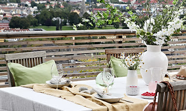 Utebord på terrasse med hvit duk, grønne puter, vaser med hvite blomster og byutsikt