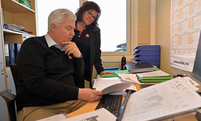 To kollegaer i svart ser gjennom dokumenter ved skrivebord med grønne permer.