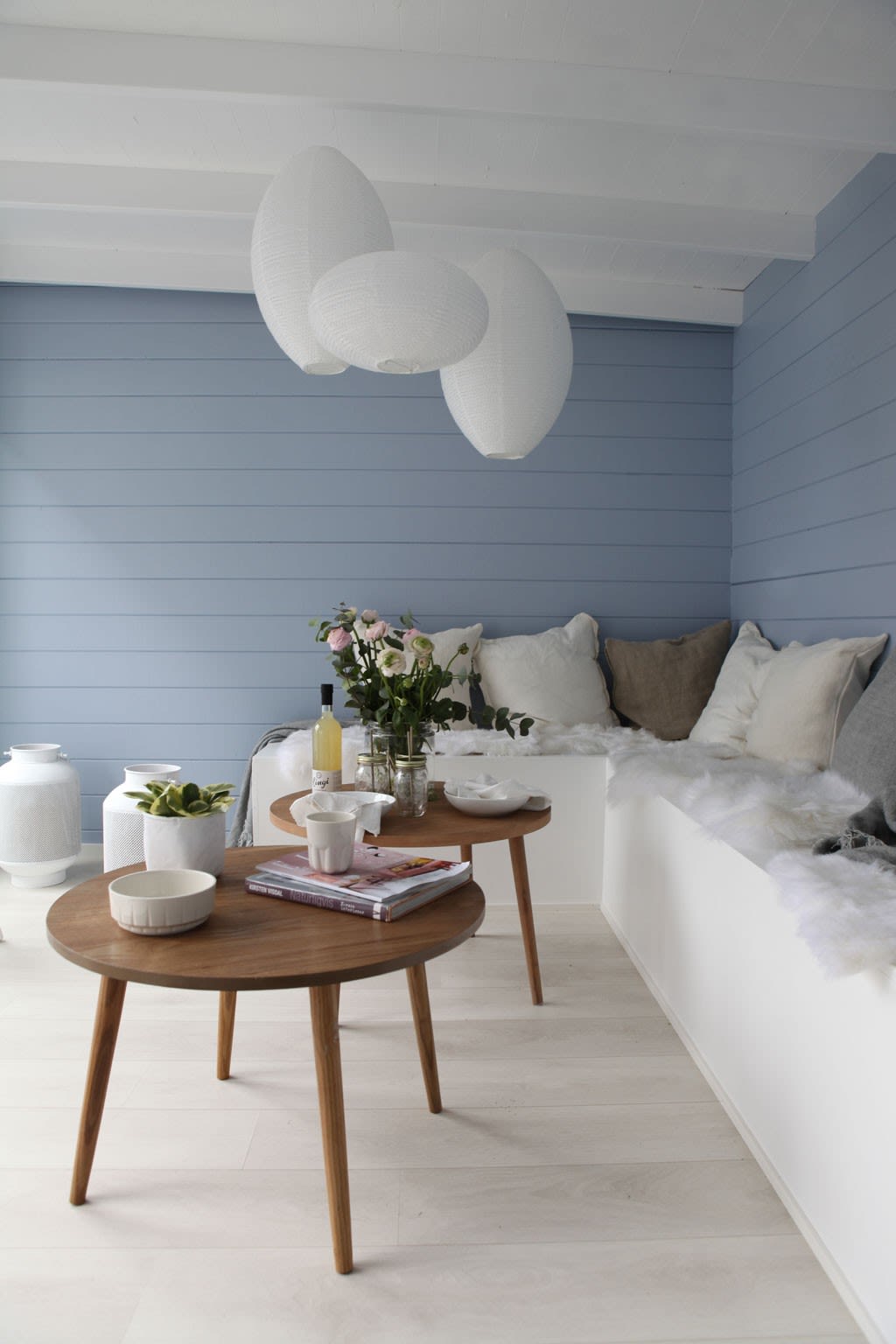 Lyst hjørne med blå panelvegg, hvite papirlykter i taket og to små runde trebord med blomster.