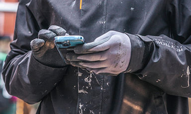 Nærbilde av maler i svart jakke med hvite malingsflekker og grå hansker som bruker mobil.