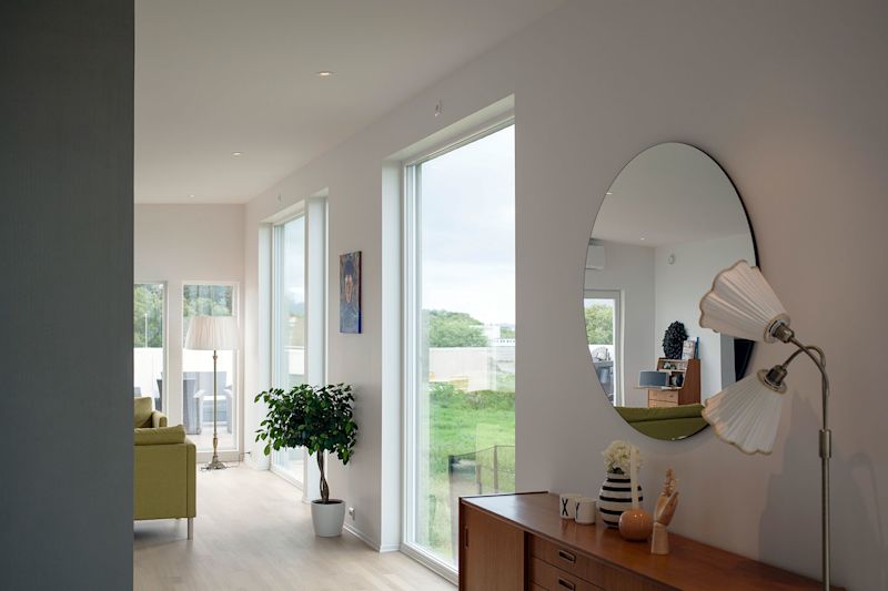 Lyst, moderne rom med store vinduer, grønn sofa, rundt speil over treskap og gulvlampe.