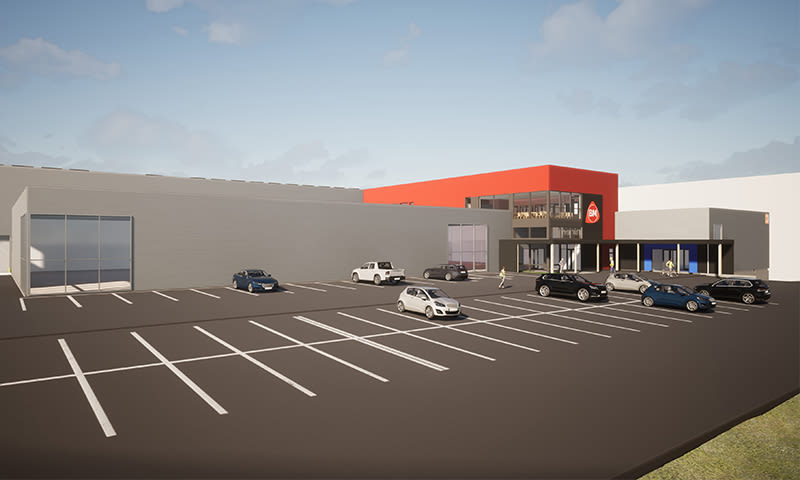 Stor grå butikkbygning med rødt inngangsparti og BM-logo, bilparkering foran under blå himmel.