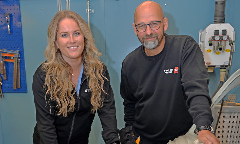 To personer i mørke gensere i blått verksted, med verktøy på veggen og maskin i bakgrunnen