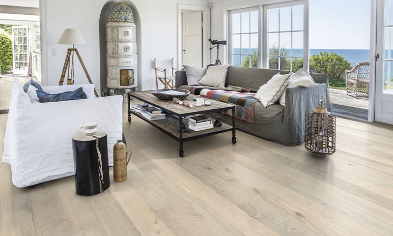 Lyst stue med hvit og grå sofa, rustikt trebord, lyst parkettgulv og havutsikt gjennom store vinduer.