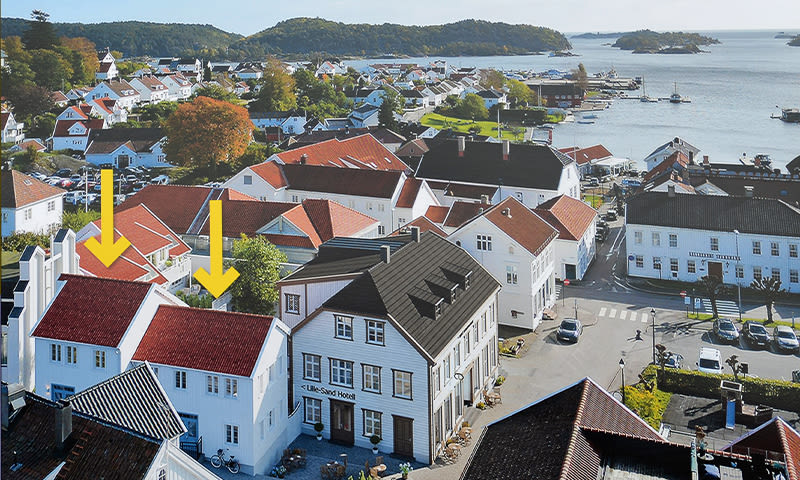 Oversiktsbilde av hvite trehus med røde tak, piler markerer to hus, fjord og holmer i bakgrunnen.