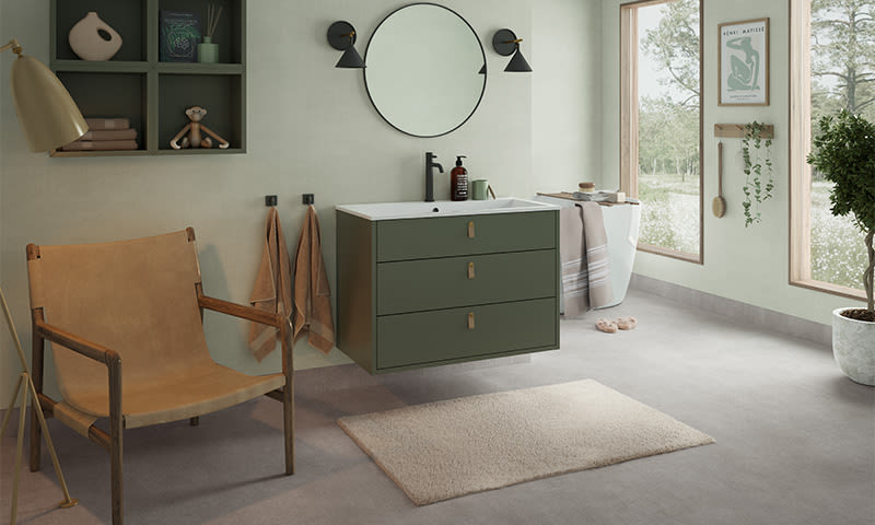 Lyst bad med grønt veggsatt servantskap, rundt speil, brun skinnstol, vindu og plante