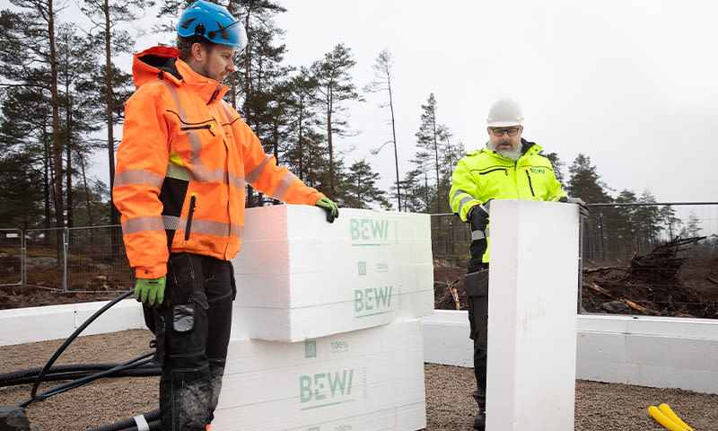 To byggarbeidere i oransje og gul refleksjakke stabler hvite BEWI-isoblokker på byggeplass.