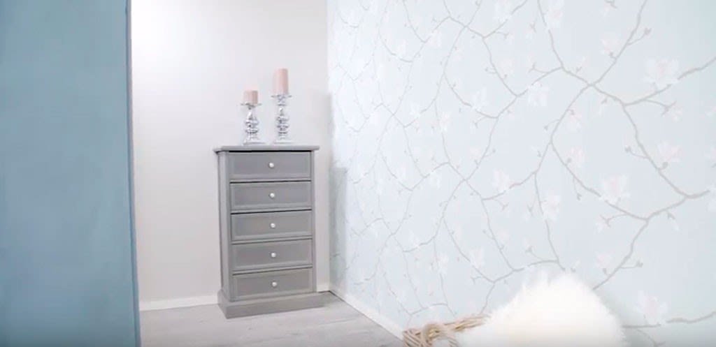 Lysegrå kommode med fem skuffer og rosa lys på toppen, foran blomstrete tapet og hvit pelskurv.