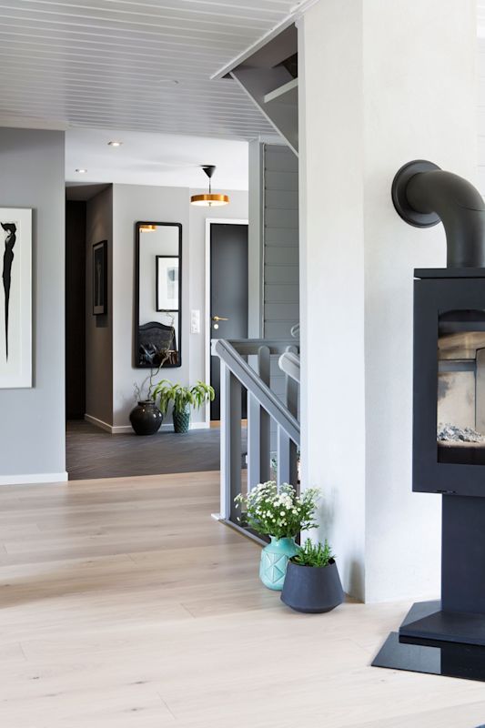 Moderne lys hall med grå vegger, speil, svart vedovn, lyse tregulv og grønne planter.