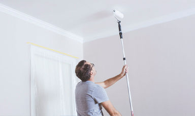 Mann i grå t-skjorte maler hvitt tak med lang rullestang i lyst oppholdsrom.