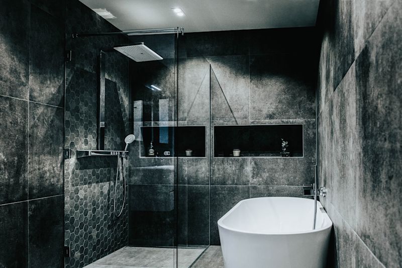 Moderne grått flislagt bad med glassdusj, innebygde hyller og hvitt frittstående badekar.