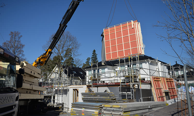 På byggeplass løfter kran rødt prefab-element over hvitt hus med stillas under klar blå himmel.