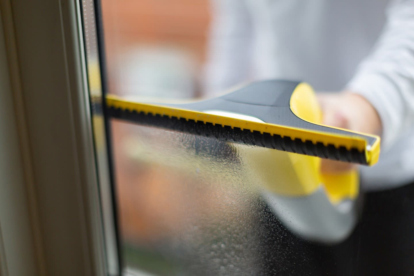 Hånd med gul-svart gummiskrape fjerner vann fra vindu
