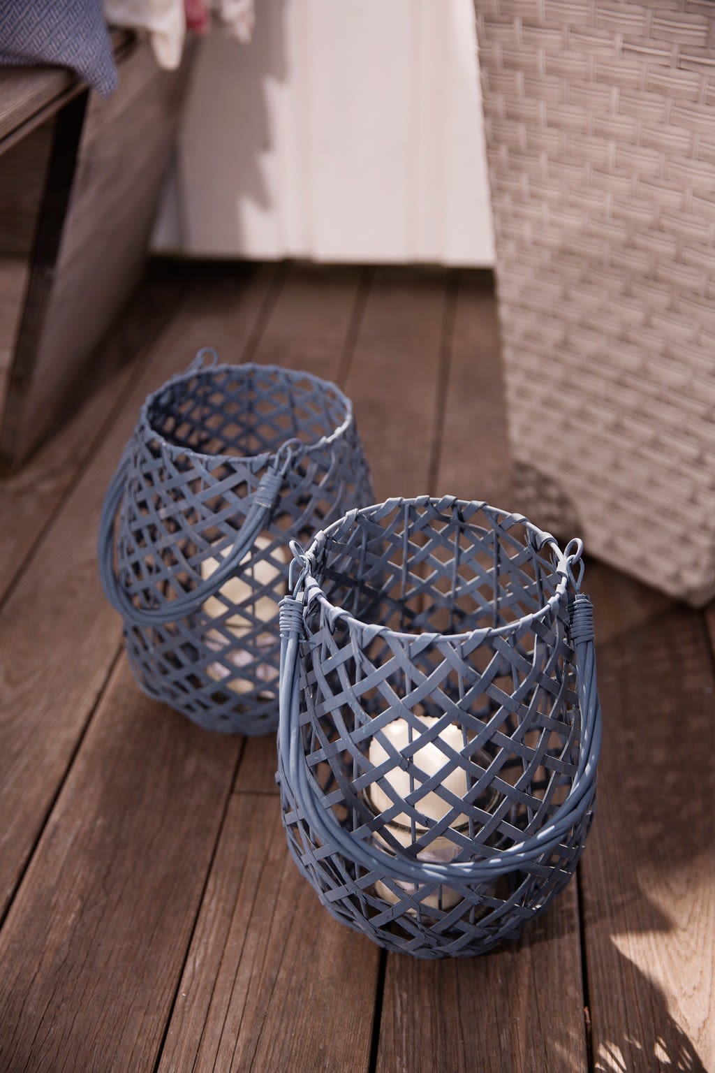 To blå flette-lykter med hvite lys på mørkt tregulv ved hvitt rottingmøbel.