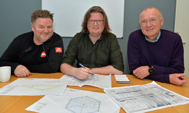 Tre personer ved lyst bord foran blågrå vegg med tekniske tegninger, en skriver med penn.